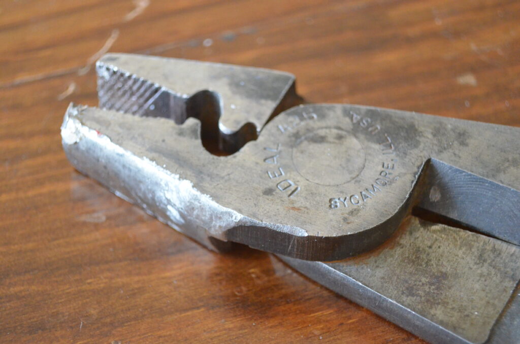 Closeup of a pair of crimping pliers used for electrical work. From left to right, the pliers show a traditional pliers end, a crimping portion, the hinge, with text saying it was mad by a US company, and wire cutters. The pliers have been placed on a very worn wooden surface.