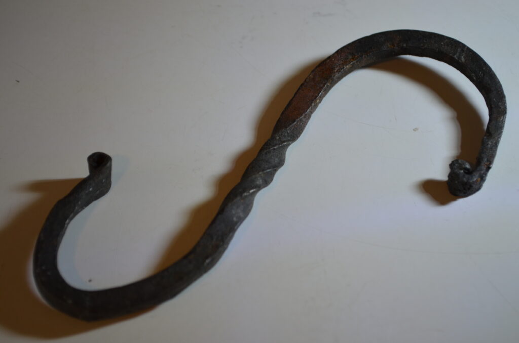 An iron hook in the shape of an S with decorative twists in the center and curls on the ends. This tool was simple enough for a beginner blacksmith to make but also incredibly useful for hanging items like lanterns. In the 90s my Explorer Post made several of these while doing historical reenacting and I still have use for mine every now and then.