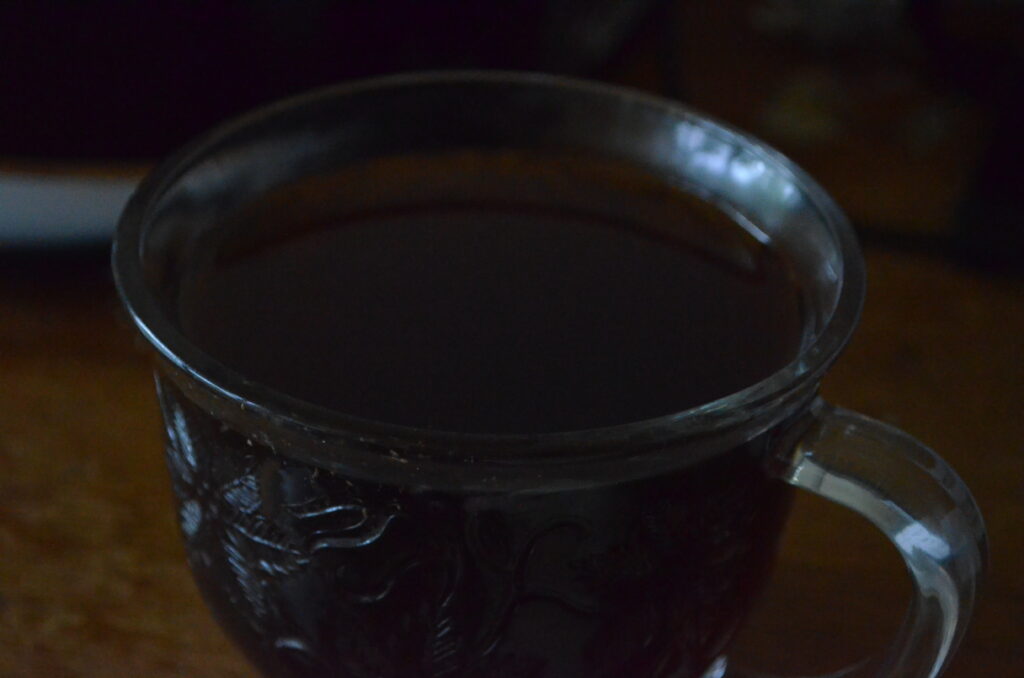 A glass cup meant for tea or coffee sits on a wooden surface. It is filled to near the top with a dark liquid. While there is enough light for the photo, the scene is relatively dark as no lights have been turned on this early in the morning.