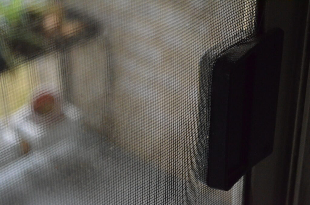 View through the screen of my patio door. The clasp is in focus (dust and all), while bricks, flower pots, and plants are blurry in the background due to a shallow depth of field.