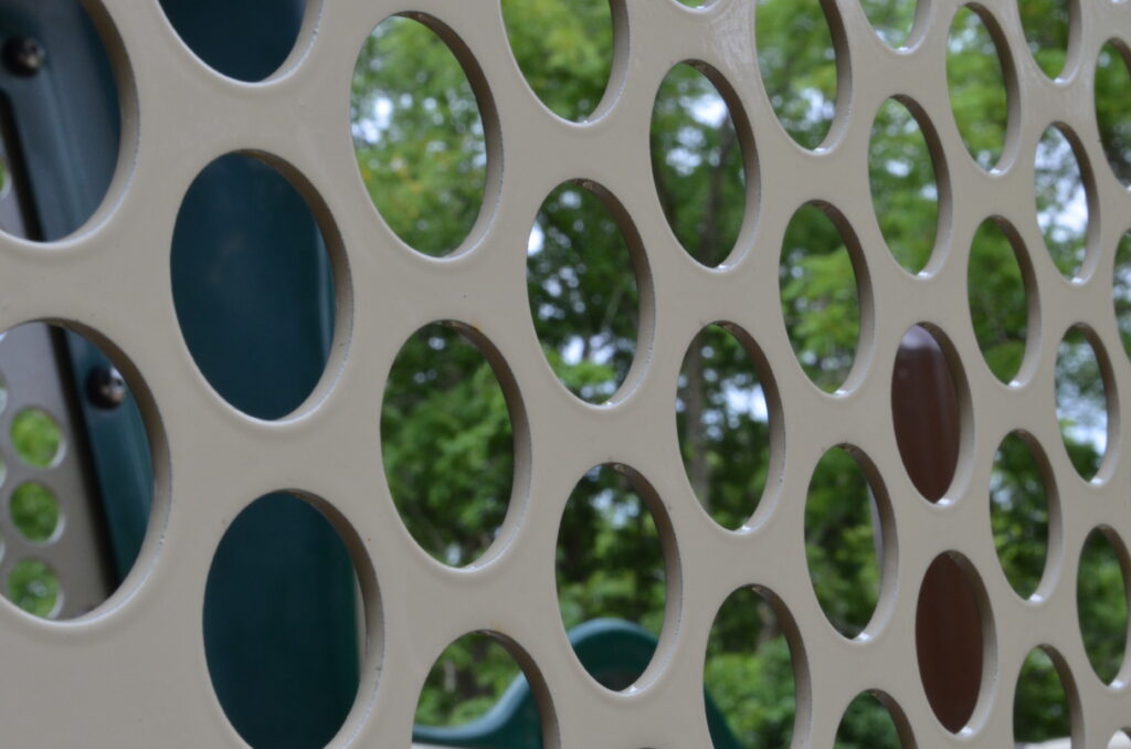 Alt Text: Close-up of a safety barrier on a playground slide. It is tan with a pattern of circles cut out of it. Trees can be seen in the background through the holes.