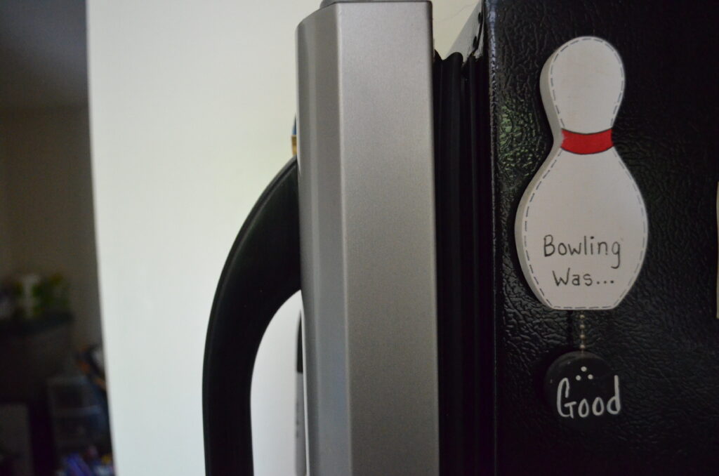 Alt Text: A wooden fridge magnet painted to look like a bowling pin. A wooden circle painted to look like a bowling ball hangs from the pin by the chain. On the pin are the words "Bowling Was..." while the ball has the word "Good" on one side and "Bad" on the other.