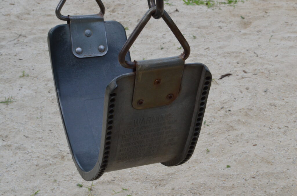 Alt Text: A rubber seat playground swing over sand. The warning label on the underside is just barely visible.