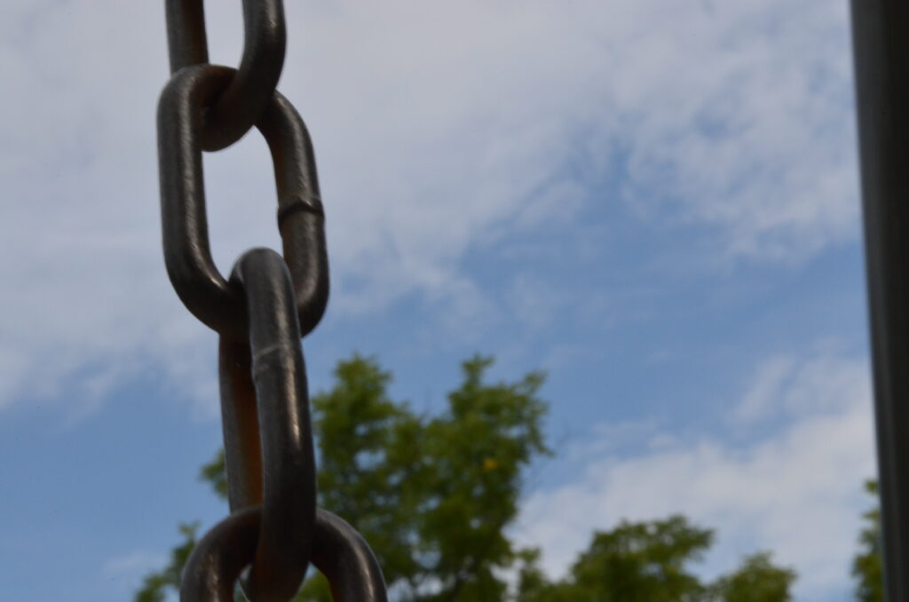 A playground swing chain, painted black, is at a slight angle on the left side of the photo. On the right is part of the support structure for the swing. Most of the image is a cloudy sky and treetops.