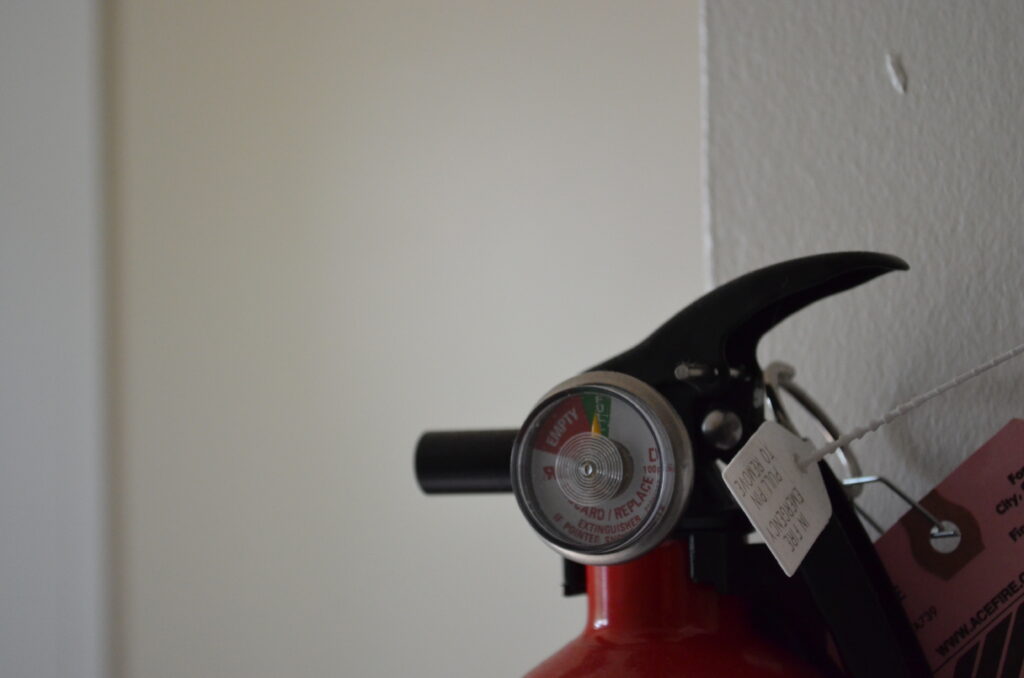 Alt Text: The top end of a small fire extinguisher in front of white walls, most of which are out of focus due to a very shallow depth of field.