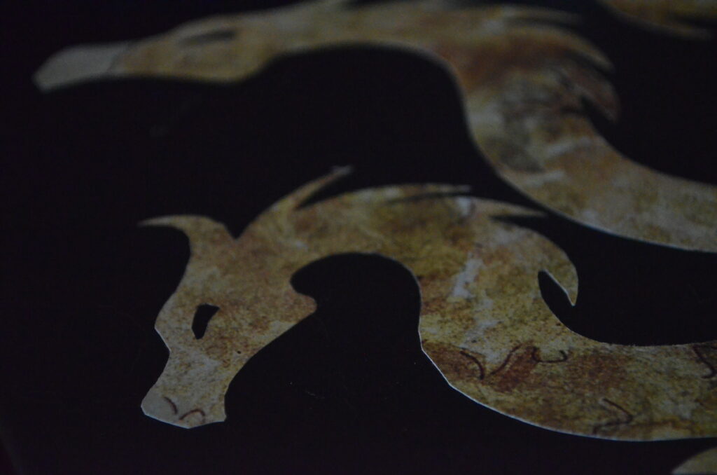 Alt Text: A pair of dragon heads cut out of fancy looking scrapbook paper on a black surface. There is a very low depth of field so the one on the top of the image is far enough away to be out of focus.