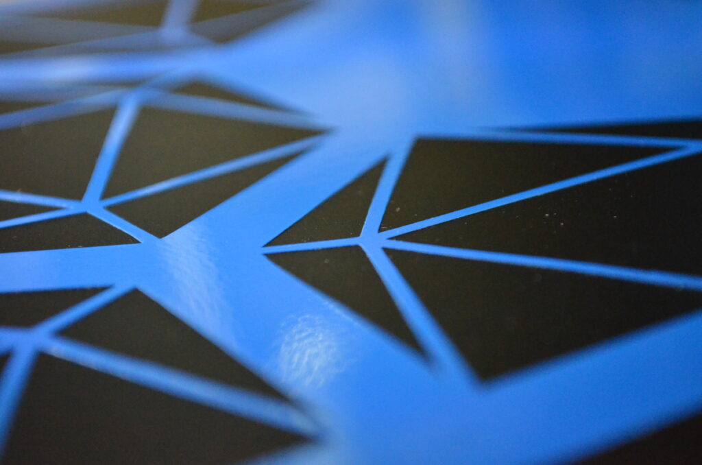 Alt Text: Blue vinyl with triangles cut out of it to form a pattern that resembles 20 sided dice. A black background is seen through the triangular holes.