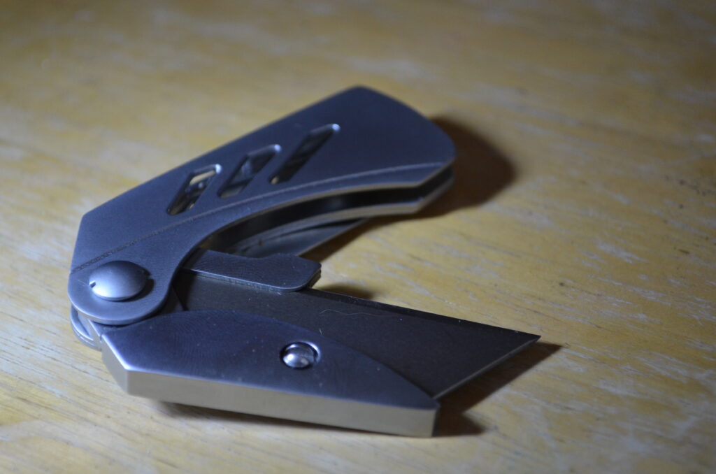 Alt Text: A stainless steel utility knife is partially opened on a wooden surface. There is a strong light source off camera to the left causing some darker shadows.