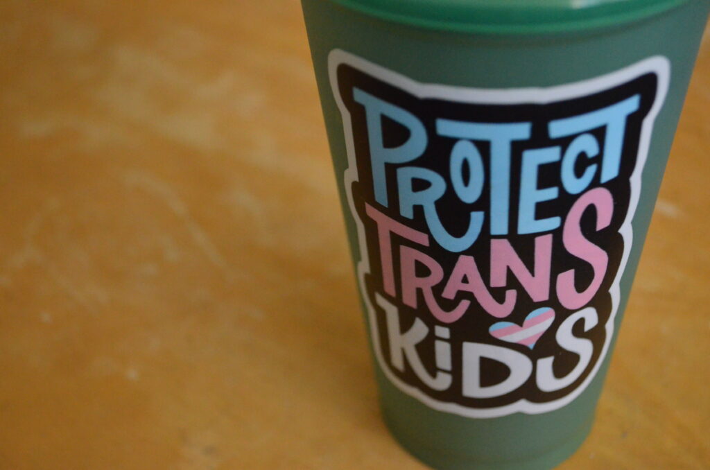 Alt Text: A green, plastic coffee travel mug that was a gift from a student sits atop a worn, wooden surface. The Starbucks logo has been covered up after the fact by a large sticker that says "Protect Trans Kids." I work in a school system where this is not something that will get me in trouble.