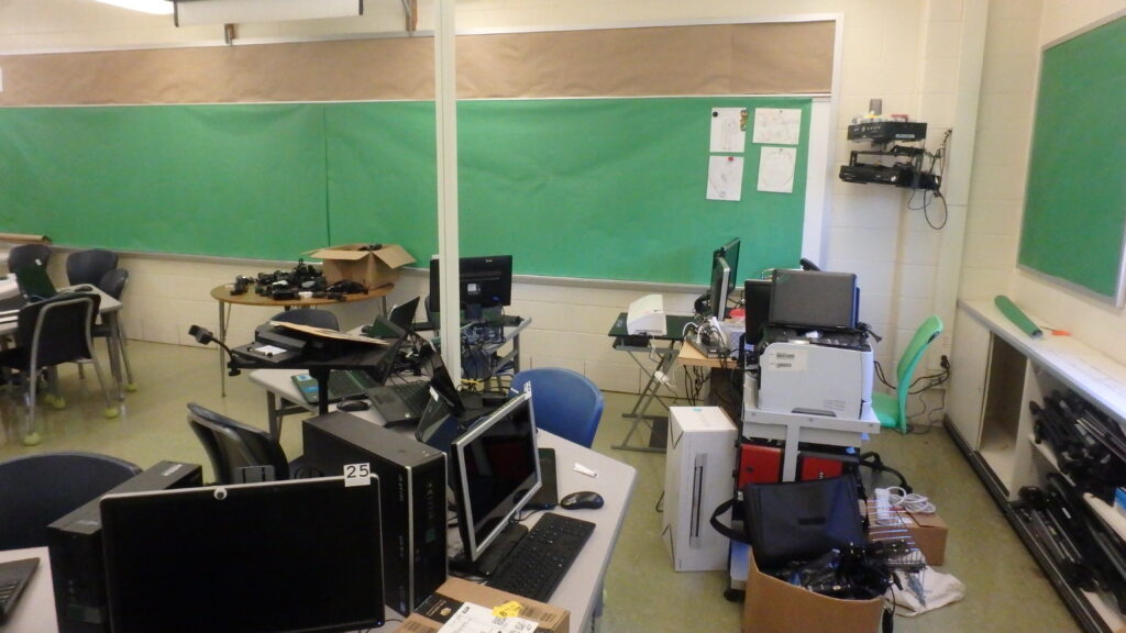 Alt Text: Photo of a computer lab that looks just as cluttered, if not more, as the last photo of the computer lab. Now, however, the whiteboards and bulletin boards are covered with green paper for chroma key projects. A selection of cameras are charging in the back of the room.